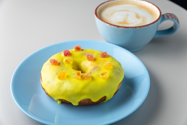 Donut covered with yellow glaze and multicolored marmalade on blue plate and cup of cappuccino