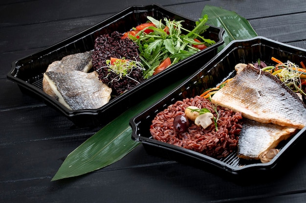 Dorado fillet with brown rice and pickled vegetables in containers on a black wooden background Takeaway Diet and healthy food