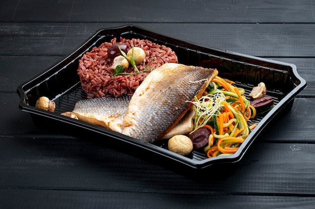 Dorado fillet with brown rice and pickled vegetables in containers on a black wooden background Takeaway Diet and healthy food