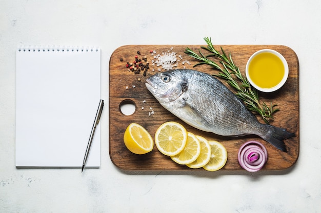 Dorado fish on a rustic wooden cutting board with spices and a notebook for recipe or menu. Top view on a white background.