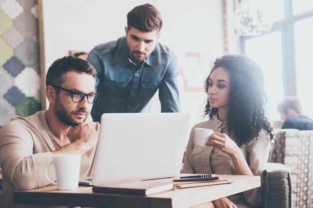 Double-checking everything. Young handsome man using laptop while sitting at the office table on business meeting with his coworkers