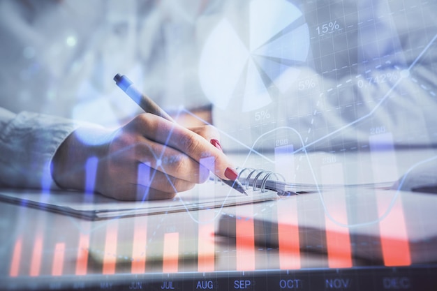 Double exposure of woman's hands making notes with forex graph hologram Concept stock market analysis