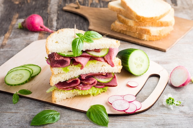 Double sandwich with pastrami and fresh vegetables and herbs on a cutting board. American snack. Rustic style