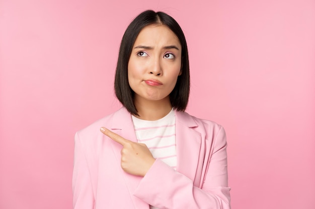 Doubtful business woman sulking and grimacing while pointing looking left at banner with skeptical face expression posing against pink background