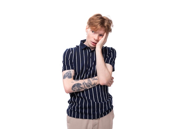 Doubtful young european blond guy in a striped polo shirt with a tattoo on a white background