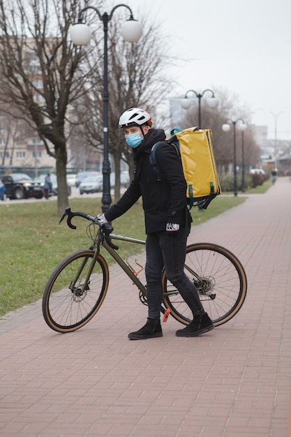 Dourier wearing medical face mask and thermo backpack, walking in the city with his bicycle