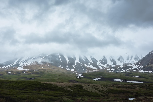 Dramatic alpine landscape with snowy mountains in gray low clouds Bleak atmospheric scenery of tundra under lead gray sky Gloomy minimalist view to mountain range among low rainy clouds in overcast