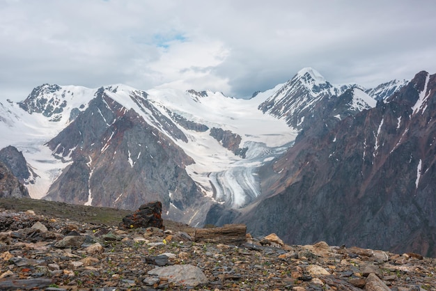 Dramatic landscape with high snowy mountain range with long glacier tongue and pointed peak in sunlight under cloudy sky Atmospheric view from stone hill to large snow mountains at changeable weather