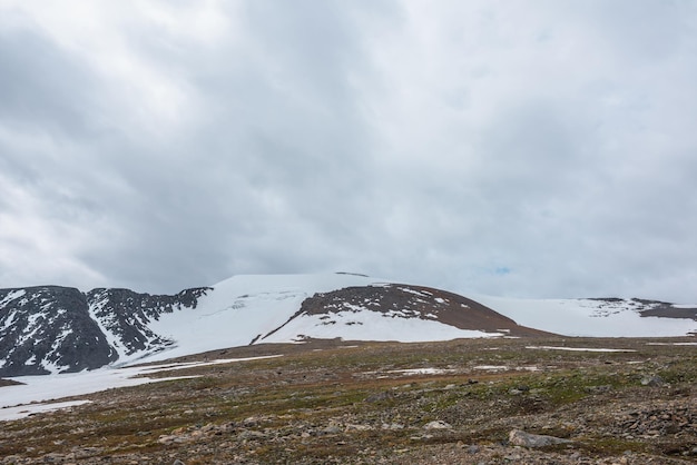 Dramatic landscape with stone field and large snowy mountain dome under gray cloudy sky Atmospheric scenery with high snow mountain in dome shape in center at cloudy weather Overcast in mountains