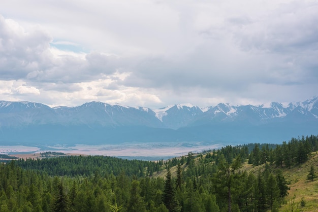 Dramatic mountain landscape with green coniferous forest hills and high snowy mountain range under rainy cloudy sky Atmospheric aerial view to conifer forest and large snow mountains in overcast