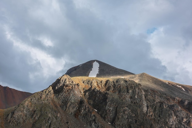 Dramatic mountain landscape with sunlit pointy peak with snow under gray cloudy sky at changeable weather Atmospheric mountain scenery with peaked top with sharp rocks in sunlight under overcast sky