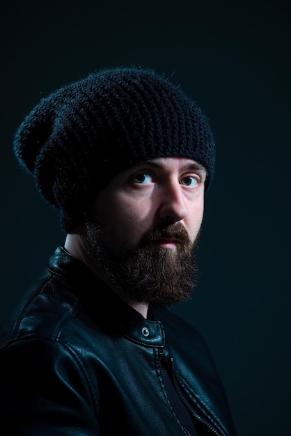 Dramatic portrait of bearded millennial guy in leather jacket on dark background.