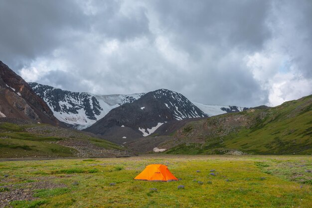 Dramatic scenery with vivid orange tent on grass in sunlit green mountain valley with view to snow mountains under gray cloudy sky Scenic landscape with snow mountain range at changeable weather