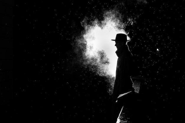 Dramatic silhouette of a dangerous man in a hat at night in the rain in the city in the old crime Noir style