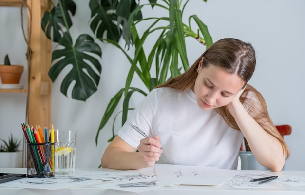 Drawings made in pencil are on the table A young woman of European appearance draws with a pencil on white paper Graphics The process of drawing with a pencil at home at a white table