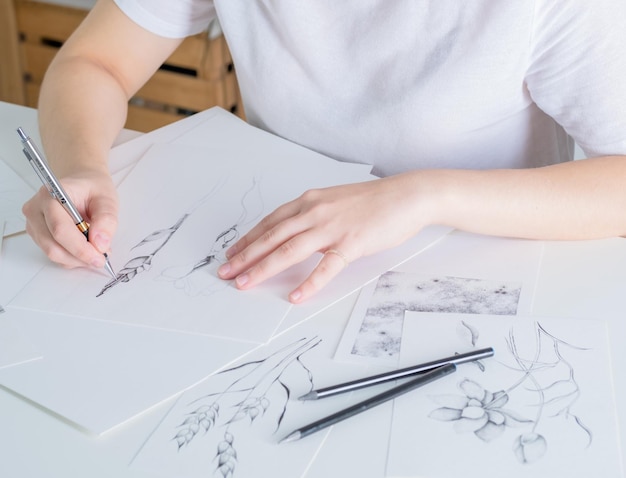 Drawings made in pencil are on the table A young woman of European appearance draws with a pencil on white paper Graphics The process of drawing with a pencil at home at a white table