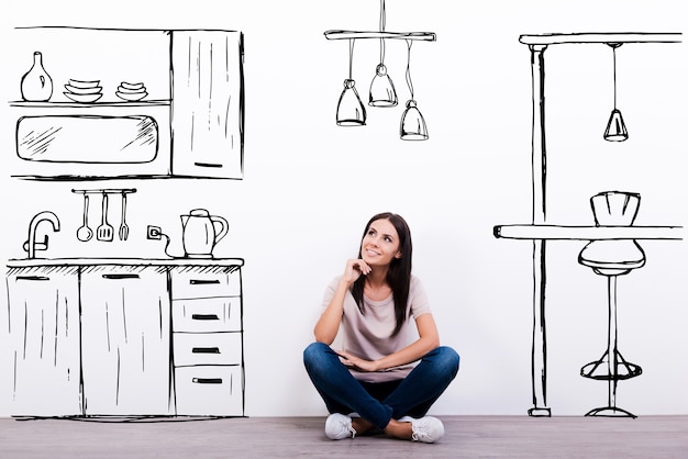 Photo dreaming about new kitchen. cheerful young woman smiling while sitting on the floor against white background with drawn kitchen