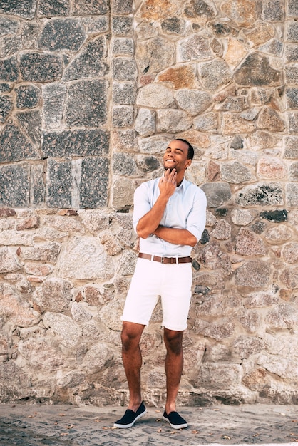 Dreaming about something good. Full length of cheerful young African man in smart casual clothes holding hand on chin and looking thoughtful while standing against the stoned wall outdoors