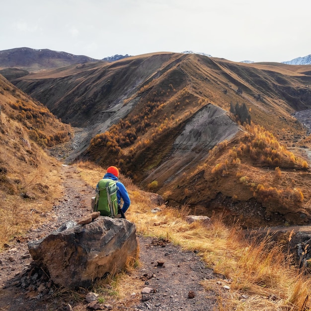 The dreaming hiker sits on a large boulder and happily looks into the distance against the background of blue dramatic clouds against the background of Caucasus mountains Rest time on a hike