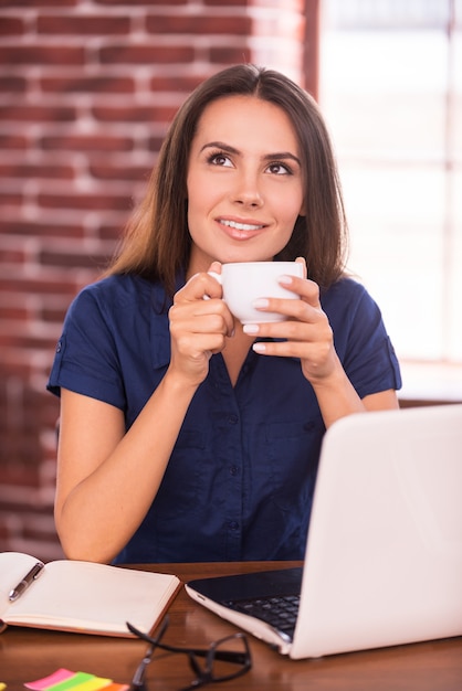 Dreaming up business ideas. Thoughtful young woman holding cup with hot drink and looking away while sitting at her working place