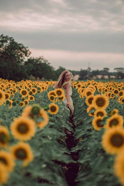 Dreaming young woman in yellow dress holding hat and walking away in a field of sunflowers at summer, view from her back. Looking back. copy space