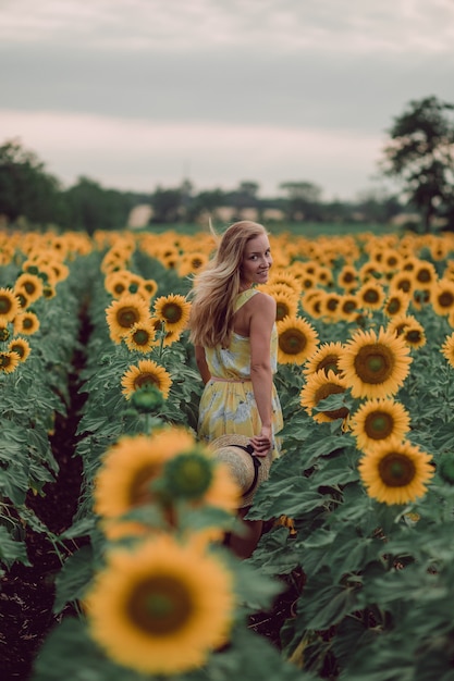 Dreaming young woman in yellow dress holding hat and walking away in a field of sunflowers at summer, view from her back. Looking to the camera. copy space