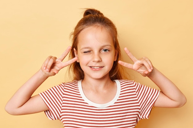 Dreamy cheerful dark haired little girl showing vsign near eyes winking wearing casual style striped Tshirt standing isolated over beige background