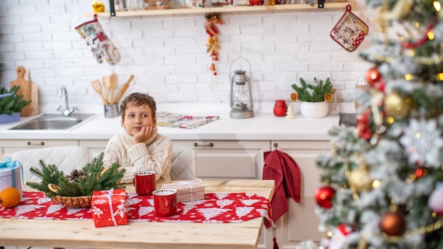 A dreamy child of 4 years in a light sweater sits at the kitchen table and looks away. in the background the bright kitchen is decorated with pine garlands