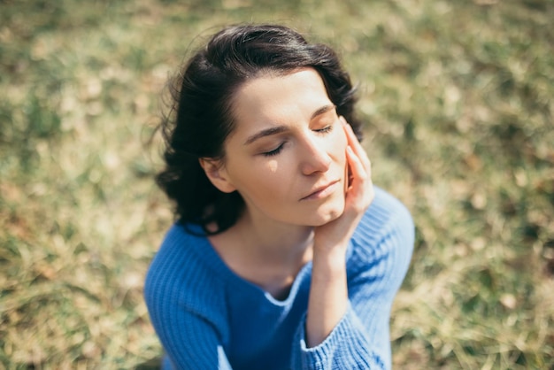 Photo dreamy female with closed eyes with hand on face portrait of attractive brunette caucasian young woman with windy hair dreaming outside on spring or on summer sitting in park lifestyle concept