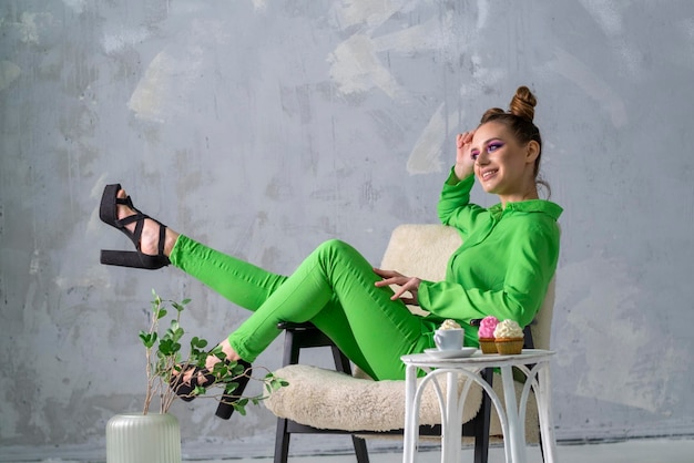 Dreamy girl poses on chair on gray wall background Young woman in green suit tastes cupcakes with cream and coffee