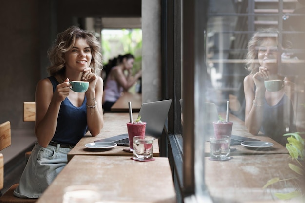 Dreamy happy lucky young alluring girl sitting coffee table cafe drink cappuccino smoothie gazing through window passersby thoughtful creating project working freelance digital nomad use laptop