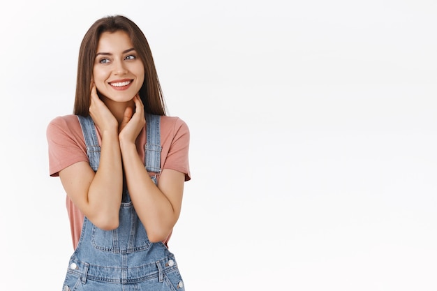 Dreamy, lovely and feminine beautiful woman in overalls, t-shirt, touching neck gently with both hands and smiling as looking away modest or shy, flirting, standing coquettish over white background