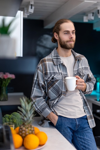 Dreamy man enjoying morning coffee standing in kitchen with modern interior drinking fresh warm beverage at home