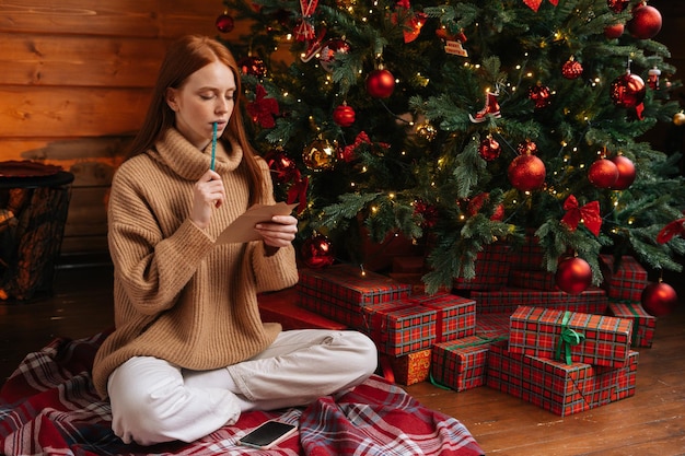 Dreamy pensive redhead young woman wearing winter sweater writing Christmas letter to Santa Claus