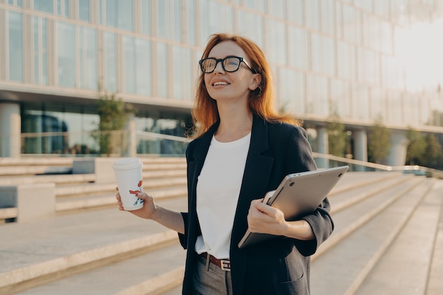 Dreamy redhead woman in formal clothes carries notepad tablet disposable cup of coffee