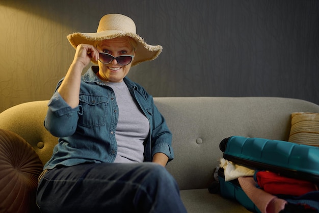 Dreamy smiling senior woman tourist sitting on sofa with packed clothes luggage for journey