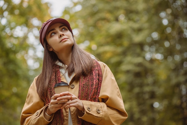 dreamy woman with paper cup of takeaway coffee standing in nature in fall