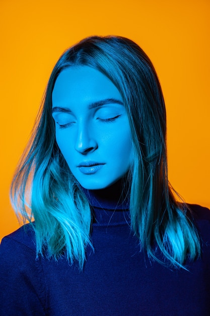 Photo dreamy young female model in turtleneck with bright neon light on face standing against orange background with closed eyes