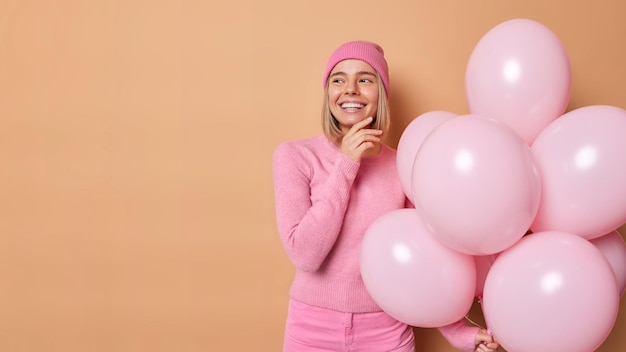 Dreamy young lovely woman organizes farewell party touches jawline gently looks away has good mood holds bunch of helium balloons poses against brown background blank copy space for promotion