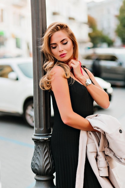 Dreamy young woman in pleated dress posing after date with eyes closed. Outdoor portrait of shapely european model in black outfit spending time on the street alone.