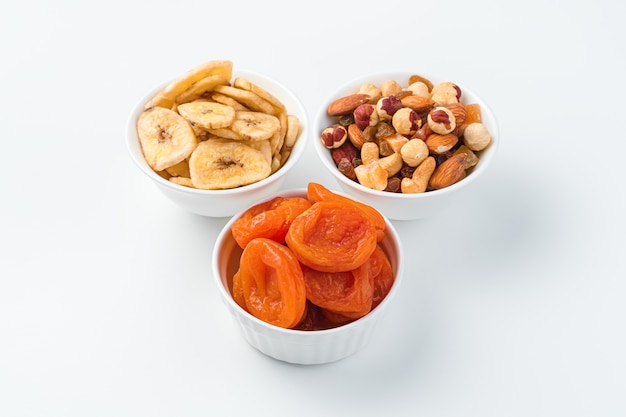 Dried apricots, banana chips and a mixture of nuts in three bowls on a white wall. Healthy snacks.