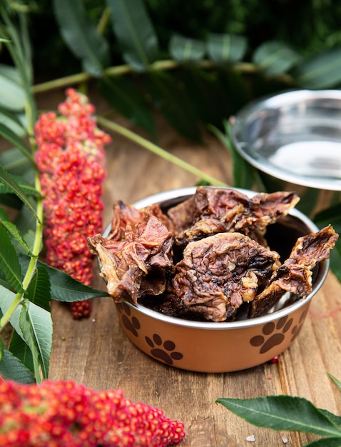 Dried beef udder in the dog vessel among the greenery on the wooden board and a silver plate with water inside on the background. Chewing treats for domestic dogs. Natural dental treats for promotion