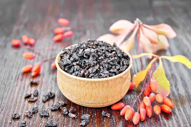 Dried black barberry in a bowl, sprigs with fresh berries and leaves on wooden board background