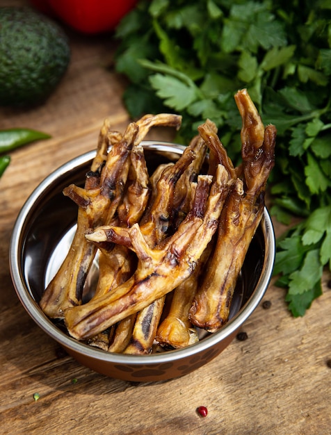 Dried chicken legs in the dog vessel among the greenery on the wooden board and some vegetables on the background. Chewing treats for domestic dogs. 