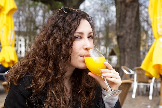 Photo drink fresh juice orange yellow girl coat curly hair smile street netherland hague sun spring brunette hand holding glass