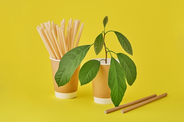 Drinking tubes made of paper and cornstarch, biodegradable material and eco paper glasses with green sprout leaves on yellow trend color 2021 background. Zero waste and plastic free concept. Top view.