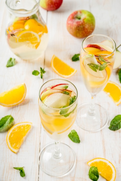 Drinks and cocktails. White autumn sangria with apples, orange, mint and white wine. In glasses for champagne, in a pitcher, on a white wooden table.