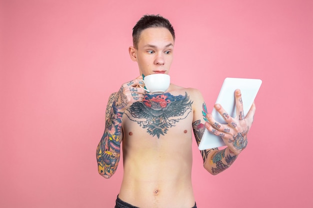 Drinks coffee, using tablet. Portrait of young man with freaky appearance on pink background.