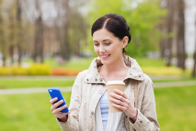 drinks, leisure, technology and people concept - smiling woman with smartphone texting message and coffee in park