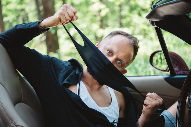 Driver in black face mask in red car open top on nature background.